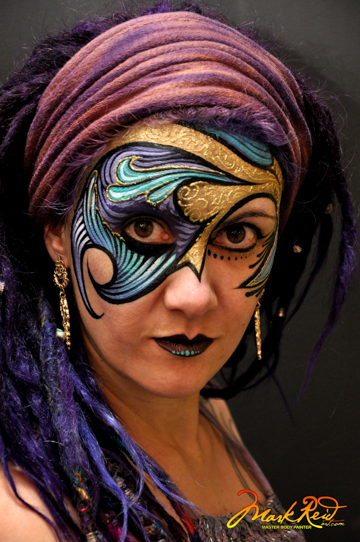 black haired woman in a painted on mask around her eyes in gold and blue that swirls
